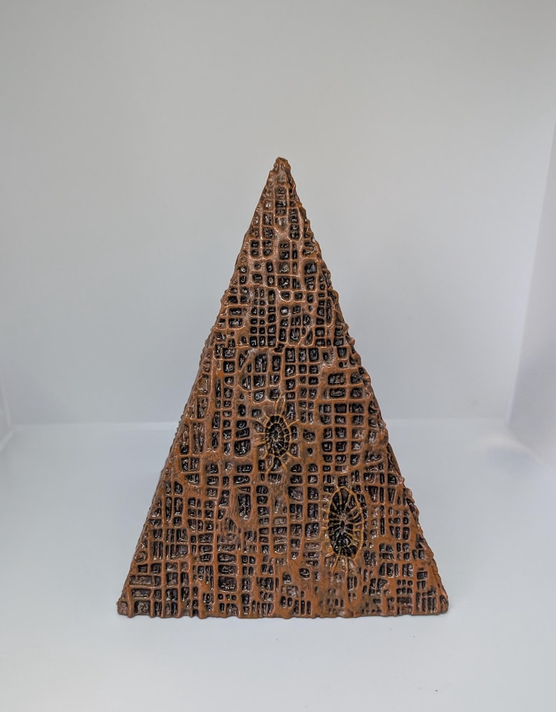 K&ouml;nnte beinhalten: Eine bronzefarbene, pyramidenf&ouml;rmige Skulptur mit einer strukturierten, gitterartigen Oberfl&auml;che. Die Skulptur weist zwei ovale, fossilartige Designs auf. Das Kunstwerk wird vor einem schlichten, wei&szlig;en Hintergrund pr&auml;sentiert.