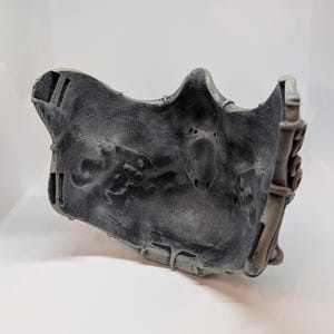 May include: A gray, textured mask with intricate details. The mask has a weathered appearance with a dark gray base and lighter gray highlights. The design includes sculpted elements and a contoured shape, suggesting a fantasy or cosplay theme.