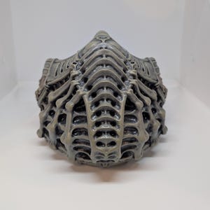 May include: A gray, intricately designed face mask with a skeletal, biomechanical appearance. The mask features a series of layered, ribbed structures and numerous small openings, creating a textured, three-dimensional effect. The overall aesthetic suggests a futuristic or science fiction theme.