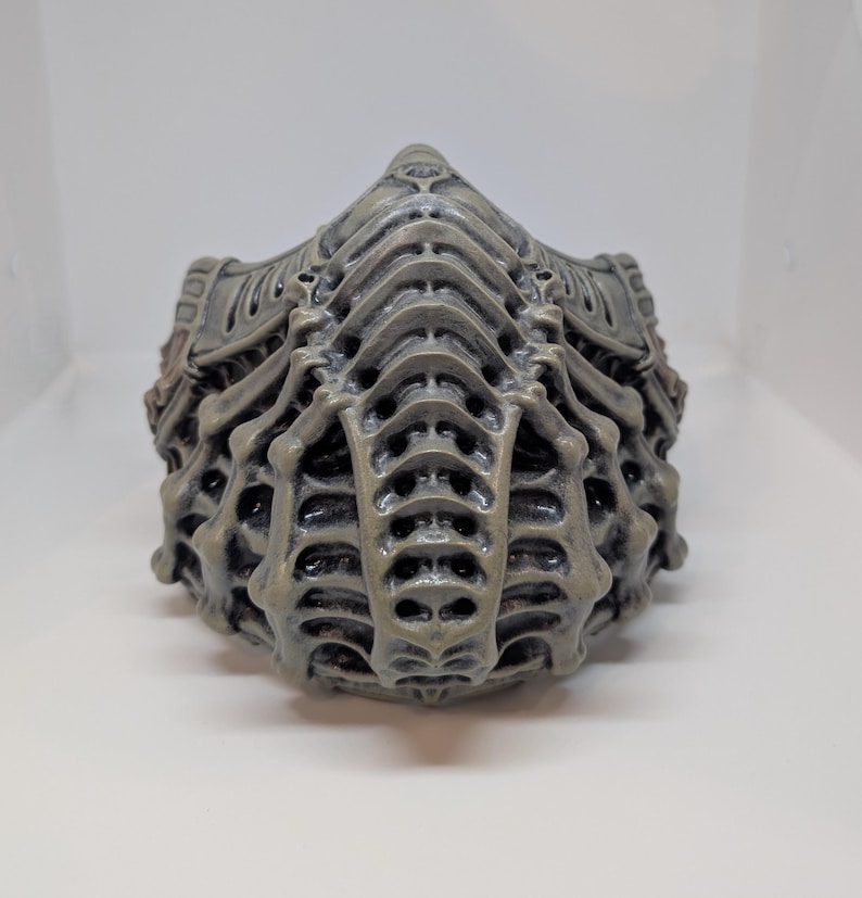 May include: A gray, intricately designed face mask with a skeletal, biomechanical appearance. The mask features a series of layered, ribbed structures and numerous small openings, creating a textured, three-dimensional effect. The overall aesthetic suggests a futuristic or science fiction theme.