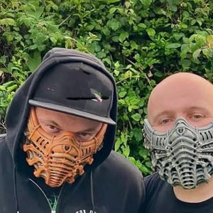May include: Two people wearing intricately designed face masks. One mask is orange, the other is gray. The masks have a detailed, mechanical appearance, with layered patterns and a futuristic aesthetic. The background is a dense wall of green foliage.