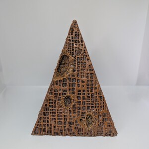 K&ouml;nnte beinhalten: Eine braune, pyramidenf&ouml;rmige Skulptur mit strukturierter Oberfl&auml;che und einem gitterartigen Muster. Die Skulptur weist kreisf&ouml;rmige, fossilartige Designs auf. Die Farbe ist ein warmer, erdiger Ton, der eine nat&uuml;rliche oder gealterte &Auml;sthetik suggeriert. Etwa 15 cm hoch.