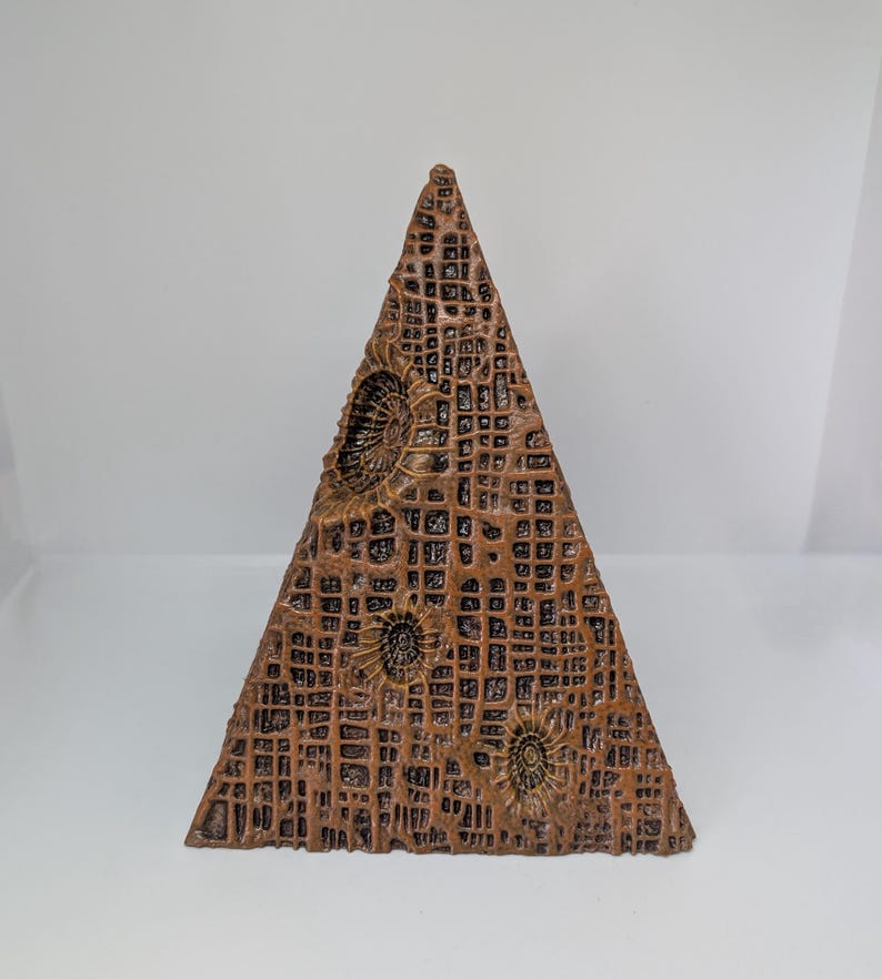 K&ouml;nnte beinhalten: Eine braune, pyramidenf&ouml;rmige Skulptur mit strukturierter Oberfl&auml;che und einem gitterartigen Muster. Die Skulptur weist kreisf&ouml;rmige, fossilartige Designs auf. Die Farbe ist ein warmer, erdiger Ton, der eine nat&uuml;rliche oder gealterte &Auml;sthetik suggeriert. Etwa 15 cm hoch.