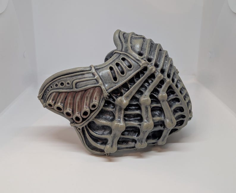 May include: A gray and brown mask with a skeletal design. The mask has a textured surface with a series of holes and a ribbed structure. The mask is made of a hard material and is likely a costume piece.