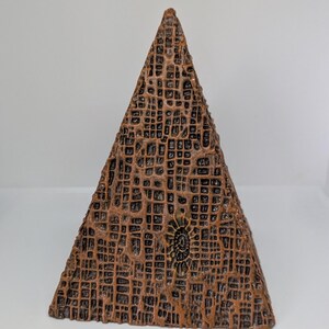 K&ouml;nnte beinhalten: Eine braune, pyramidenf&ouml;rmige Skulptur mit einer strukturierten, gitterartigen Oberfl&auml;che. Die Skulptur hat ein kleines, kreisf&ouml;rmiges Design auf einer Seite. Die Farbe ist eine Mischung aus Braun und Bronze.