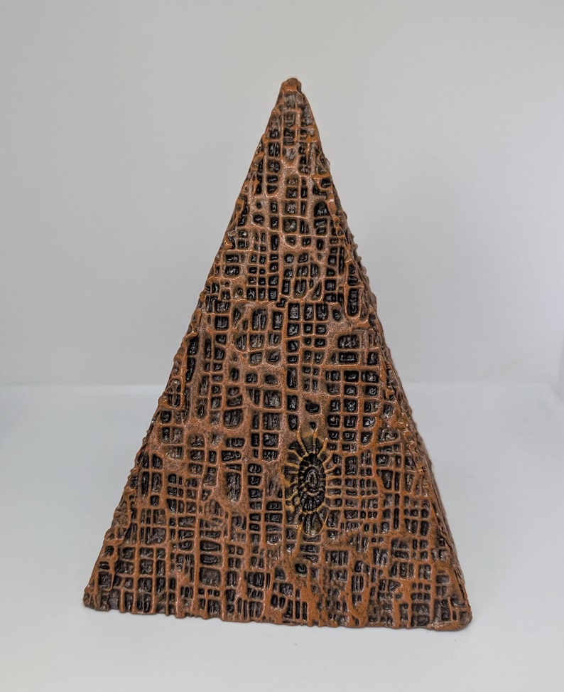 K&ouml;nnte beinhalten: Eine braune, pyramidenf&ouml;rmige Skulptur mit einer strukturierten, gitterartigen Oberfl&auml;che. Die Skulptur hat ein kleines, kreisf&ouml;rmiges Design auf einer Seite. Die Farbe ist eine Mischung aus Braun und Bronze.