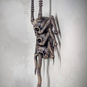 Op de afbeelding: Een donkere, gedetailleerde sculptuur van een wezen bestaande uit opgestapelde schedels, insectachtige poten en een gesegmenteerde staart. De sculptuur heeft een verweerde, bronsachtige afwerking en is op een lichtgekleurde muur gemonteerd.