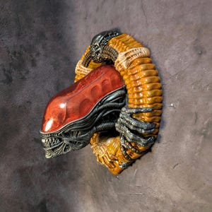 May include: A detailed sculpture of an Alien Xenomorph head and body, featuring a red, metallic head with sharp teeth and a ribbed, orange body. The sculpture has a textured, organic appearance with metallic accents, showcasing a blend of colors and intricate details.