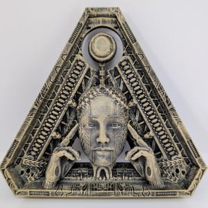 May include: A beige and gray triangular wall sculpture featuring a stylized human face with intricate patterns. The face is framed by a geometric design with mechanical elements, and the hands are positioned near the ears.