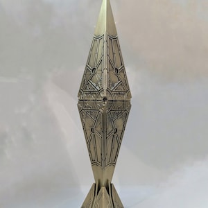 Puede incluir: Una escultura metálica alta, con forma de diamante y una parte superior puntiaguda. La escultura tiene intrincados patrones geométricos y se apoya sobre una base de tres patas. El color es un dorado apagado.