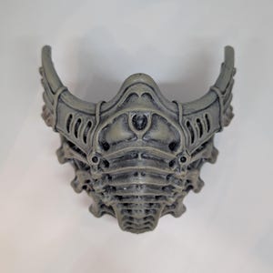 May include: A gray, intricately designed mask with a textured surface. The mask features curved horns and detailed patterns, suggesting a fantasy or sci-fi theme. The mask is made of a durable material.