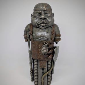 May include: A detailed sculpture of a figure with a weathered, metallic appearance. The figure wears a headpiece, leather straps, and a chest piece with a grenade. It holds a rifle and a shield, with a cylindrical lower body.