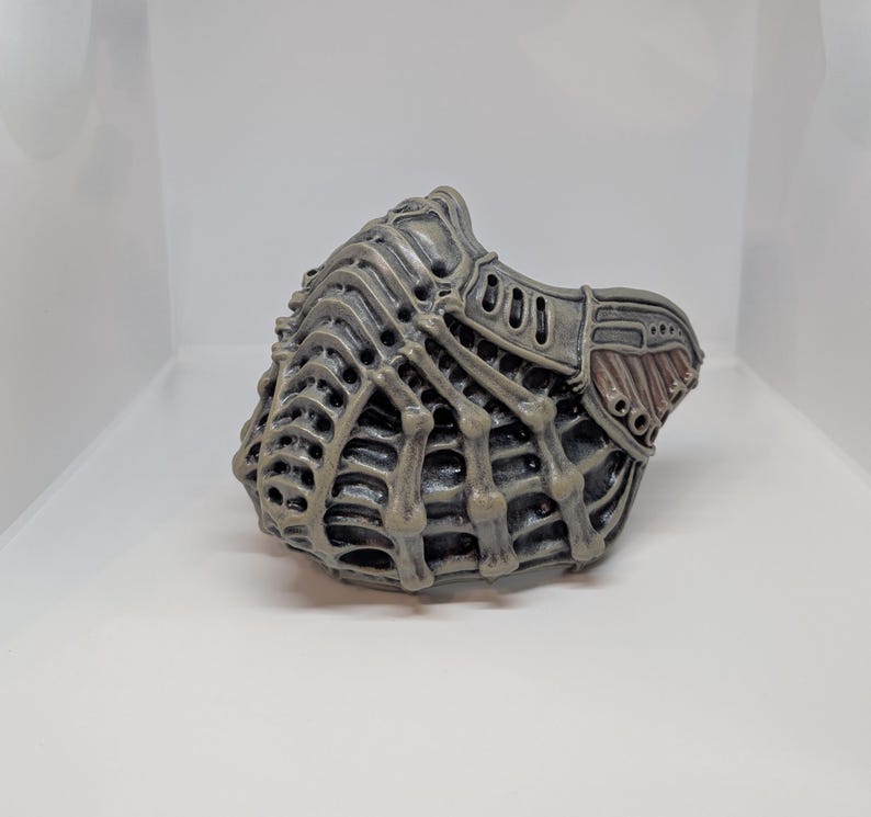 May include: A gray, intricately designed mask with a bio-mechanical aesthetic. The mask features a complex network of ribbed structures and openings, with a darker, textured section on the side. The overall design suggests a futuristic or science fiction theme.