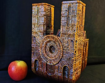 Könnte beinhalten: Eine detaillierte, bronzefarbene Skulptur einer Kathedrale mit zwei Türmen und einem kreisförmigen Fenster. Ein rot-gelber Apfel liegt neben der Skulptur. Die Skulptur hat komplizierte drahtartige Details.
