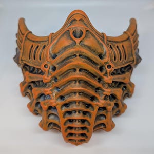 May include: An orange and brown mask with a detailed, textured design. The mask has a complex, layered structure with horn-like protrusions on the sides. The color scheme is a blend of orange and brown, creating a weathered appearance. The mask appears to be a costume or decorative item.