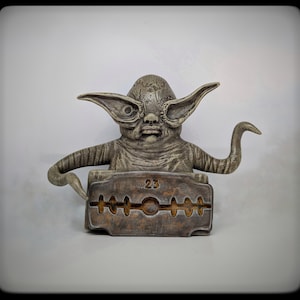 May include: A weathered, gray-toned sculpture of a creature with large ears and a stylized face. The figure is positioned above a rusty-brown, blade-shaped base with the number "23" inscribed. The sculpture has a unique, aged appearance.