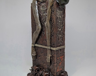Könnte beinhalten: Eine braune Skulptur mit einer skelettierten Figur, die über einer strukturierten rechteckigen Säule drapiert und mit einem Seil befestigt ist. Die Figur trägt einen dunkelgrünen Helm. Die Basis besteht aus einem Haufen Schädel, der Buchstabe Z ist rot.