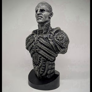 May include: A detailed bust sculpture of a humanoid figure with intricate, biomechanical detailing. The sculpture is primarily silver-gray, mounted on a black base. The figure's face has a stern expression, and the design suggests a science fiction or fantasy theme.