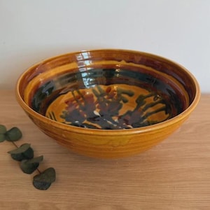 May include: A round ceramic bowl with a glossy mustard yellow exterior and a wide rim. The interior features a dark brown and black glaze with a hand-painted abstract floral pattern in deep red and dark green tones, creating a layered effect.