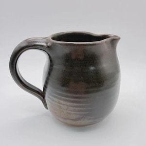 May include: A dark brown ceramic pitcher with a rounded body, a curved handle on the left side, and a small pouring spout. The surface features subtle horizontal ridges around the lower section and a smooth, glossy finish throughout.