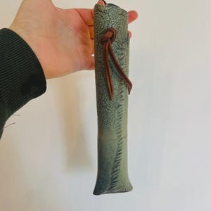 May include: A long, cylindrical, sage green leather case with a textured, crackled surface finish. The case features a brown leather cord tied in a knot near the top opening, providing a secure closure for the slender, vertical storage pouch.