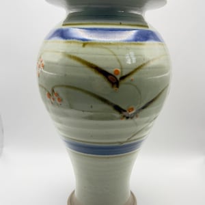 May include: A ceramic vase with a pale green glaze, featuring horizontal blue bands around the neck and lower body. The surface is decorated with dark brown vine-like patterns and small orange dots, with a wide rim and a flared base.
