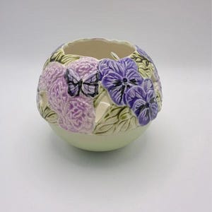 May include: A spherical ceramic candle holder featuring a light green base and a textured upper section decorated with raised purple pansy flowers, green leaves, and a single black and white butterfly. The piece has several cut-out openings.