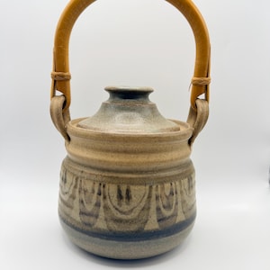 May include: A ceramic teapot with a speckled beige and grey glaze featuring a dark blue decorative pattern around the base. The vessel has a rounded body, a matching lid, and a curved, light brown bamboo handle attached to two side loops.