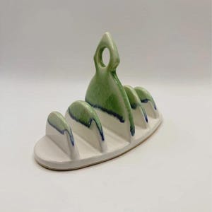 May include: A ceramic toast rack with a white base and four slots. The top edges of the dividers and the central handle are glazed in a gradient of light green and dark blue, featuring a smooth, glossy finish and a central circular handle.