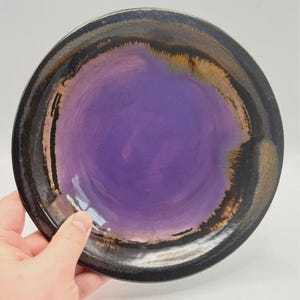 May include: A round ceramic plate featuring a vibrant purple centre that transitions into a dark, glossy black rim. The glaze shows subtle brown and metallic-toned highlights where the colours blend, creating a smooth, reflective surface.