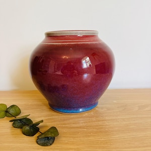 May include: A round ceramic vase with a glossy finish, featuring a deep red color that transitions into a vibrant blue at the base. The vase has a wide, curved body, a narrow neck, and a light gray rim. A small sprig of dried green leaves lies nearby.