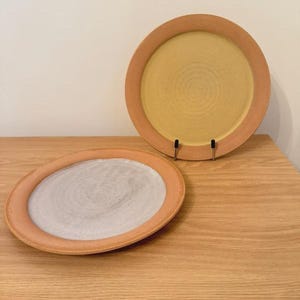 May include: Two round ceramic plates with wide, unglazed terracotta-colored rims. One plate stands upright on a small black metal stand and features a yellow center, while the other lies flat and features a light gray center with visible texture.
