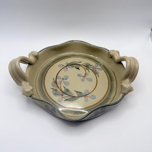 May include: A round ceramic serving dish with two ornate, curved handles on opposite sides. The dish features a cream-colored interior with a hand-painted floral motif of blue petals and green leaves arranged in a circular pattern, accented by a brown ring.