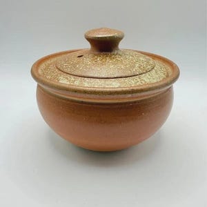 Könnte beinhalten: Eine runde, terrakottafarbene Keramikschale mit passendem Deckel. Die Schale hat eine glatte, einfarbige rotbraune Basis, während der Deckel eine texturierte, gesprenkelte beige-braune Oberfläche mit einem kleinen zentralen Knauf aufweist.