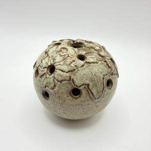 May include: A spherical ceramic flower frog with a speckled off-white glaze and a raised brown floral relief pattern. The surface features multiple circular openings distributed across the top and sides for arranging stems.