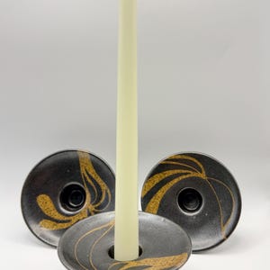 May include: Three black ceramic candle holders with a wide, circular base and a central opening. Each holder features a speckled gold-toned leaf pattern. A single, tall, off-white taper candle is inserted into the centre of the front holder.