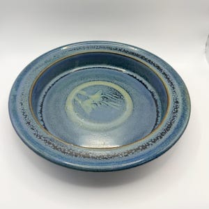 May include: A round ceramic bowl featuring a deep blue glaze with a mottled texture. The rim is accented with a dark brown border, and the centre displays a lighter, circular pattern with a subtle, abstract brushstroke design in pale yellow.