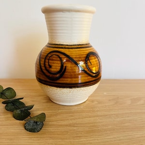 May include: A ceramic vase with a bulbous body and a flared rim. The upper neck and base are finished in a matte off-white glaze, while the center features a glossy amber-brown band decorated with two prominent, hand-painted black spiral motifs.