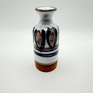 May include: A ceramic vase with a flared rim and a cylindrical body. The surface features a white glaze with dark blue and brown painted petal-like motifs, horizontal blue stripes, and a solid unglazed terracotta-colored base.