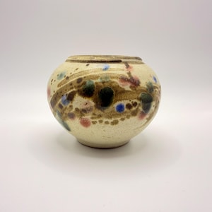 May include: A round ceramic vase with a cream-coloured base featuring a horizontal band of mottled brown, blue, and pink spots. The vessel has a wide, rounded body and a short, narrow neck with a smooth, slightly textured finish throughout.