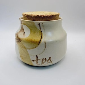 May include: A cream-coloured ceramic canister with a wide base and a tapered neck, topped with a round cork lid. The exterior features a hand-painted abstract yellow and brown brushstroke design and the word tea written in brown cursive script.
