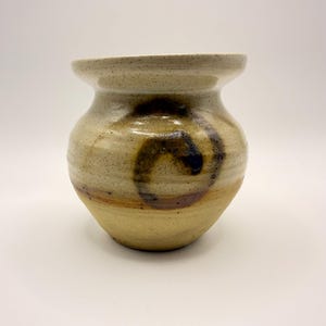 May include: A speckled ceramic vase with a wide, flared rim and a rounded body. The surface features a cream-colored glaze with brown speckles and a prominent, dark brown hand-painted spiral design encircling the center of the vessel.