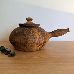May include: A round, brown clay teapot with a speckled dark brown texture. The teapot features a side-mounted handle, a flat lid with a small knob, and a wide, low-profile body. The surface has a matte finish with visible dark brown spots.