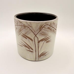 Puede incluir: Una maceta cilíndrica de cerámica con un exterior de color blanco roto moteado que presenta un diseño de hoja de palmera grabado en marrón oscuro. El interior del recipiente tiene un acabado en esmalte marrón oscuro sólido y uniforme.