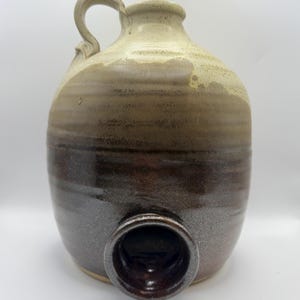 May include: A ceramic jug with a handle at the top and a circular opening near the base. The vessel features a two-tone glaze, with a creamy off-white colour on the upper portion transitioning into a deep, dark brown finish on the lower section.