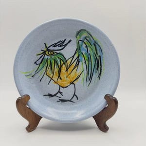 May include: A round, light blue speckled ceramic plate featuring a hand-painted illustration of a cockerel. The cockerel is rendered in yellow, green, and black brushstrokes with a stylised, expressive design. The plate is displayed on a dark wood stand.