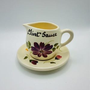 May include: A cream-coloured ceramic sauce boat with a matching saucer, featuring a hand-painted purple flower design and green leaves. The words Mint Sauce are written in black cursive script on the front of the pitcher, which has a handle.