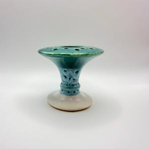 May include: A ceramic flower frog with a wide, flat top featuring multiple circular openings. The top is a glossy teal, transitioning to a speckled blue and white midsection, and ending in a solid, smooth, off-white circular base.