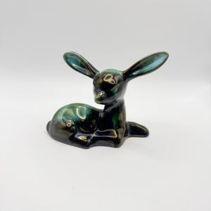 May include: A ceramic figurine of a resting deer with long ears, featuring a glossy glaze in shades of deep black and dark teal. The deer is depicted in a seated position with its head turned slightly to the side, showcasing a smooth finish.