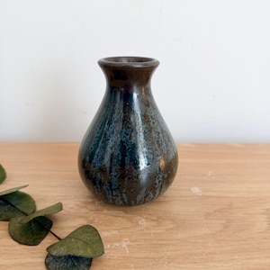 May include: A small, bulbous ceramic vase with a narrow neck and a flared rim. The surface features a dark, glossy glaze with mottled streaks of deep blue, black, and brown, creating a textured appearance across the rounded body of the vessel.